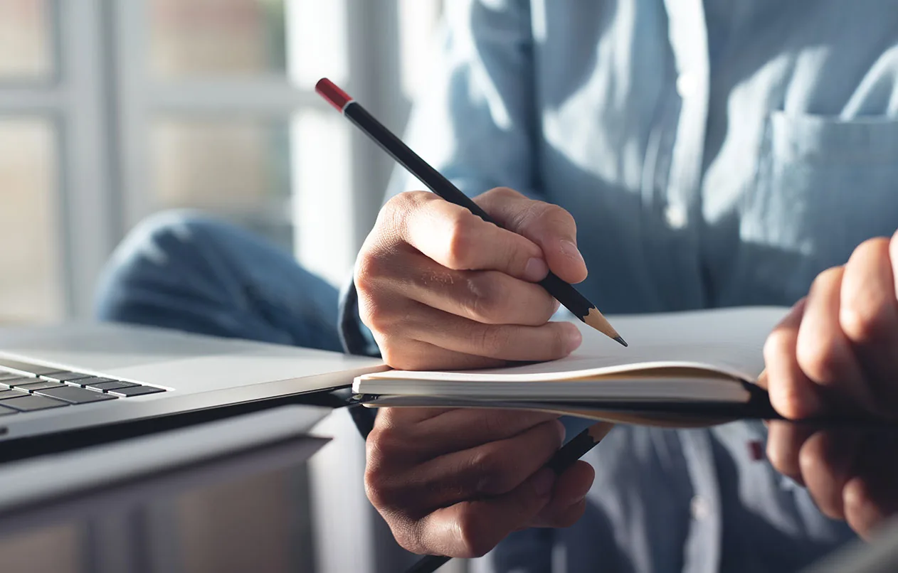 Hand mit Bleistift notiert Stichpunkte in ein Notizbuch neben dem Laptop